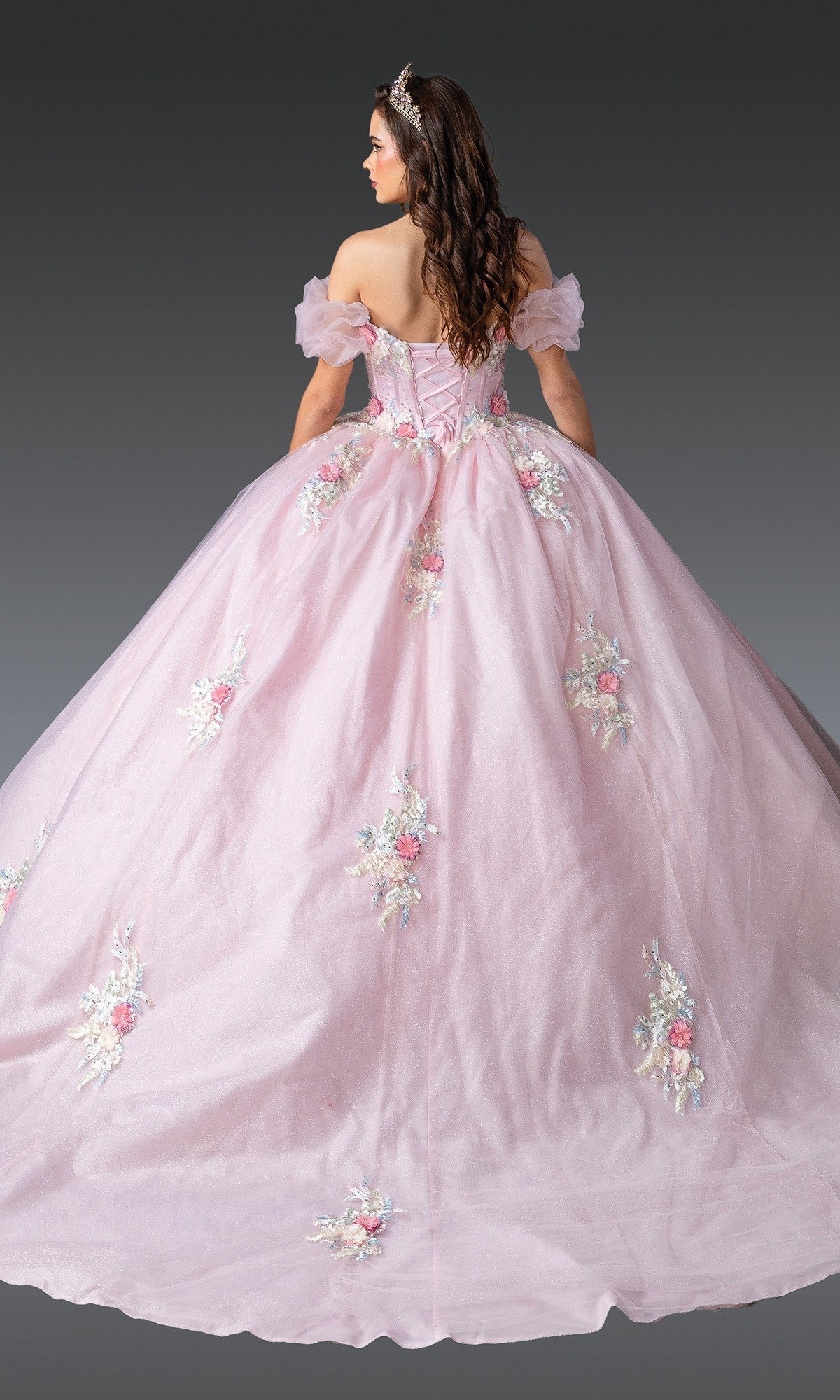 Wearing the Quinceanera Dress 1900 By Dancing Queen, a woman stands facing away and showcases the gown’s voluminous pastel pink tulle ballgown skirt and elegant corset-style back adorned with intricate 3D floral appliques.